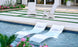 The Outdoor Escape In-Pool Furniture Bundle by Ledge Lounger features two white Signature Chaise lounge chairs with water jets for relaxation and two Adirondack chairs on a stone patio with grass accents, all bordered by pink flowers and greenery.
