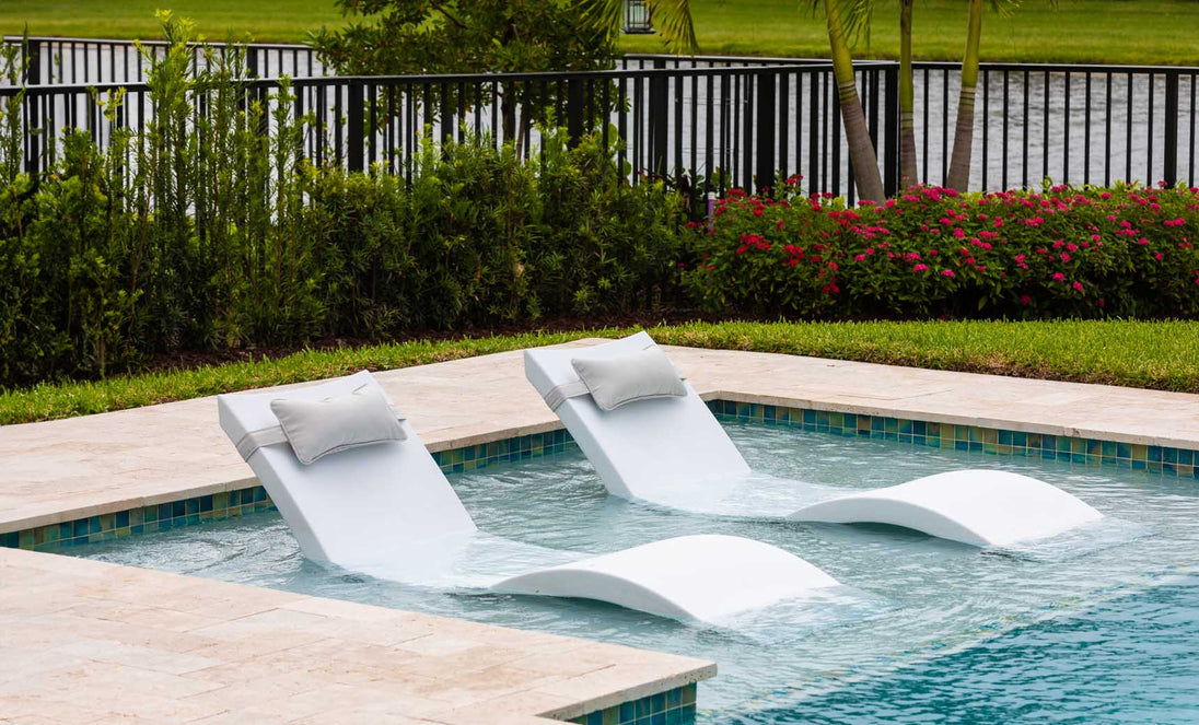 Two white chaise lounges from the Ledge Loungers Outdoor Escape In-Pool Furniture Bundle sit partly submerged in a shallow pool, facing a metal fence adorned with lush greenery and red flowers. A serene body of water and trees extend into the horizon under a clear sky.