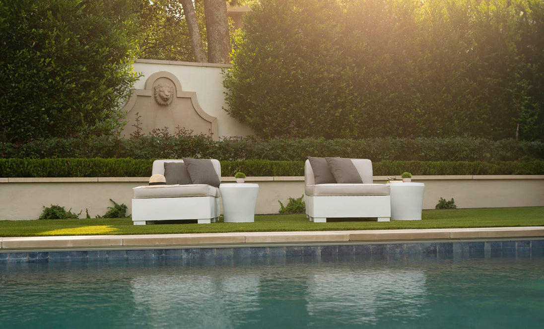 The clear poolside setting features two Ledge Loungers Affinity 2 Piece Sun Chairs in white with gray cushions. A straw hat rests on one, while tall hedges and a stone wall bask in sunlight, perfect for showcasing the UV20 rated outdoor furniture.