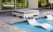 An outdoor area showcases an Affinity Square Sunbed with a Flat Cushion from Ledge Lounger, accompanied by two white lounge chairs partially submerged in the pool. A cushioned square seating area sits nearby on the gray-tiled patio, while large windows reflect greenery.