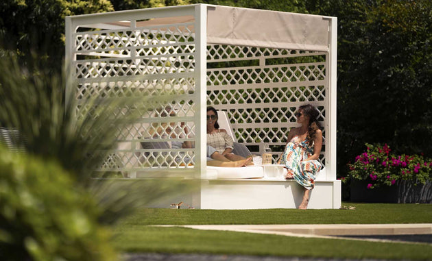 Two people relax in a durable outdoor cabana surrounded by greenery. One lounges on Ledge Loungers Shift Daybed in sunglasses, while the other, wearing a floral dress, perches on the edge, enjoying the garden view.