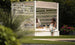 Two people relax in a durable outdoor cabana surrounded by greenery. One lounges on Ledge Loungers Shift Daybed in sunglasses, while the other, wearing a floral dress, perches on the edge, enjoying the garden view.