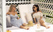 Two women in sunglasses relax on a white Shift Daybed by Ledge Lounger with a lattice backdrop, one in a blue dress and the other in plaid. A straw hat and drinks rest on the table beside them as they smile, enjoying a sunny day amidst chic outdoor decor.