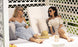 Two women in sunglasses relax on a white Shift Daybed by Ledge Lounger with a lattice backdrop, one in a blue dress and the other in plaid. A straw hat and drinks rest on the table beside them as they smile, enjoying a sunny day amidst chic outdoor decor.