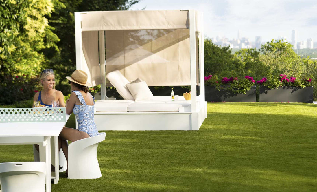 Two people sit at a white outdoor table on the grassy lawn, with a Ledge Lounger Shift Daybed nearby. One wears a sun hat and blue patterned swimsuit. Trees frame the distant city skyline, blending nature with style using durable outdoor materials.