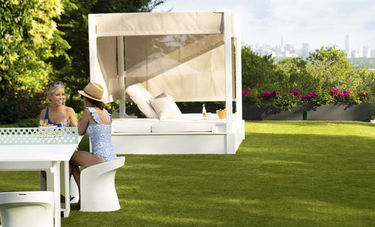 Two people sit at a white outdoor table on the grassy lawn, with a Ledge Lounger Shift Daybed nearby. One wears a sun hat and blue patterned swimsuit. Trees frame the distant city skyline, blending nature with style using durable outdoor materials.