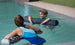 A boy and girl float in a pool using Laze Doodle cushions from Ledge Lounger. The boy smiles at the girl, who faces away from the camera. The outdoor pool is surrounded by grass and a gravel path.