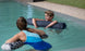 A boy and girl float in a pool using Laze Doodle cushions from Ledge Lounger. The boy smiles at the girl, who faces away from the camera. The outdoor pool is surrounded by grass and a gravel path.