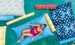 Wearing a red and pink swimsuit, someone relaxes on a large blue Laze Doodle float by Ledge Lounger in a pool. They are surrounded by colorful patterned floats featuring yellow, red, and blue designs in the clear turquoise water.
