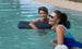 A woman in Ledge Loungers blue Laze Doodle swimsuit and sunglasses leans on the pool edge, laughing. Nearby, a smiling boy with wet hair clings to a float. The pool water is clear and theres grass in the background.