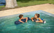 A woman and boy in the backyard pool floating on blue pool noodles.