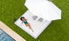 A woman in a green swimsuit relaxes on an Echo Sunbed by Ledge Lounger with gray pillows under a large white umbrella. Shes holding a phone next to a magazine and sunglasses, enjoying the tranquility of the poolside scene captured from above.