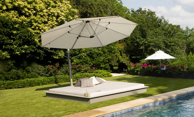A Ledge Lounger Echo Sunbed sits on a trimmed lawn by the pool, shaded by a large gray umbrella. Cushions provide comfort, and a bottle of water awaits for relaxation. In the backdrop, trees and an umbrella adorned with pink flowers add to the serene outdoor setting.