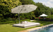 A Ledge Lounger Echo Sunbed sits on a trimmed lawn by the pool, shaded by a large gray umbrella. Cushions provide comfort, and a bottle of water awaits for relaxation. In the backdrop, trees and an umbrella adorned with pink flowers add to the serene outdoor setting.