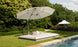 A Ledge Lounger Echo Sunbed sits on a trimmed lawn by the pool, shaded by a large gray umbrella. Cushions provide comfort, and a bottle of water awaits for relaxation. In the backdrop, trees and an umbrella adorned with pink flowers add to the serene outdoor setting.