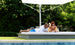 A man in a white shirt and blue shorts checks his phone, while a woman in a yellow swimsuit smiles beside him. Both relax on their Ledge Lounger Echo Sunbed by the pool under a large white umbrella, soaking up the serene outdoor ambiance surrounded by trees.