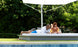 A man in a white shirt and blue shorts checks his phone, while a woman in a yellow swimsuit smiles beside him. Both relax on their Ledge Lounger Echo Sunbed by the pool under a large white umbrella, soaking up the serene outdoor ambiance surrounded by trees.
