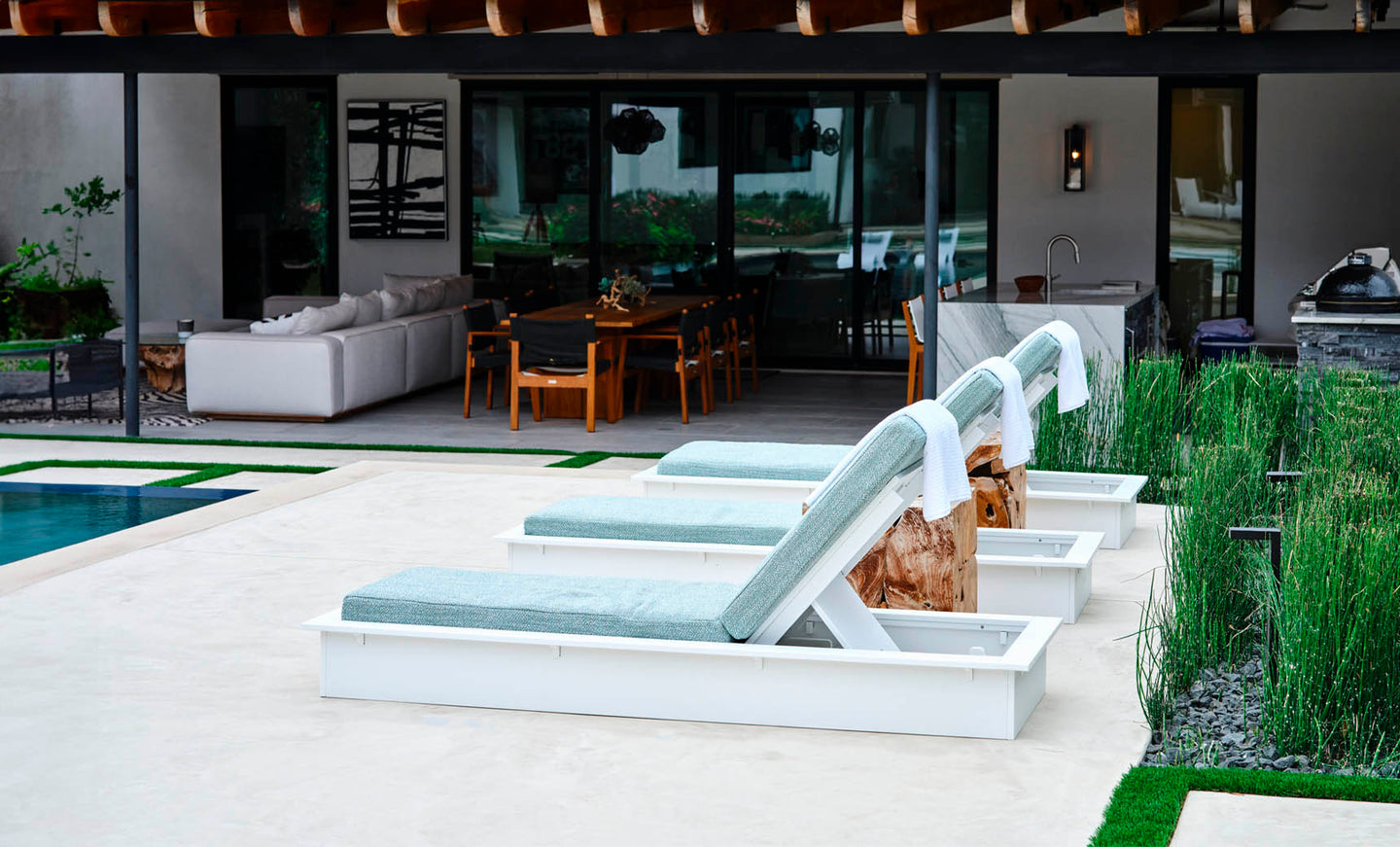 Poolside scene with three Echo Chaise loungers by Ledge Lounger, featuring light blue cushions and towels facing a shimmering swimming pool. In the background, enjoy an inviting outdoor space with comfortable sofas, a dining table, and lush greenery.