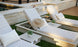 Three Echo Chaise loungers by Ledge Lounger, each with patterned cushions and a straw hat, are arranged poolside. Every adjustable chair includes a small side table with a potted plant. The area is beautifully surrounded by light stone and wooden decking.