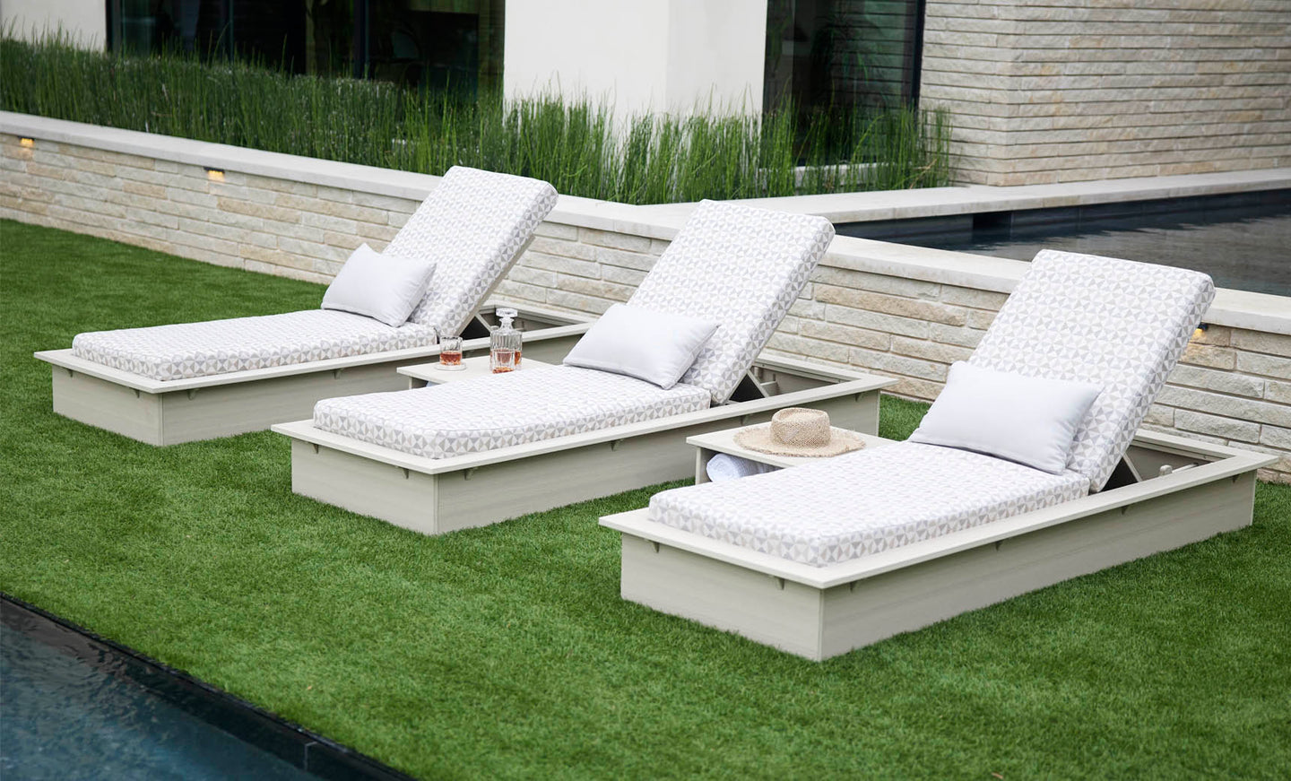 Three Echo Chaise loungers by Ledge Lounger, cushioned in white with pillows, are set on trimmed grass next to a stone-tiled pool. An adjustable chaise holds two bottles and a straw hat, enhancing the tranquil space amid lush greenery and a modern building.