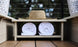An Echo Side Table by Ledge Lounger sits with a straw hat on its smooth wooden shelf, positioned above two rolled white towels. The outdoor setting features blurred greenery in the background, creating a serene atmosphere.