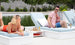 A woman in a red swimsuit relaxes on a lounge chair, smiling at a man wearing sunglasses and a pink shirt on another chair. They are by the pool with greenery around, enjoying drinks set on an Echo Side Table by Ledge Lounger.