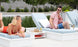 A woman in a red swimsuit relaxes on a lounge chair, smiling at a man wearing sunglasses and a pink shirt on another chair. They are by the pool with greenery around, enjoying drinks set on an Echo Side Table by Ledge Lounger.