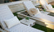 Two Ledge Lounger patterned poolside loungers with pillows sit on a lawn by the pool, one topped with a straw hat. Echo Side Tables - Final Sale, each adorned with small potted plants, are positioned between the loungers for added decor.