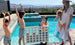 A woman in a swimsuit and sunhat holds a fluffy dog by the pool, while a young woman smiles from within it. Nearby, two children raise their arms in excitement beside Ledge Loungers Four In A Row: an oversized outdoor game with large colored chips, set against a bright sky and mountains.