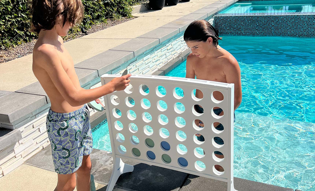Two boys in swim trunks enjoy oversized outdoor fun at the pools edge, playing Ledge Loungers Four In A Row. One places a large colored chip while the other looks on, surrounded by sunlit water and tiled edges.
