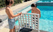Two boys in swim trunks enjoy oversized outdoor fun at the pools edge, playing Ledge Loungers Four In A Row. One places a large colored chip while the other looks on, surrounded by sunlit water and tiled edges.