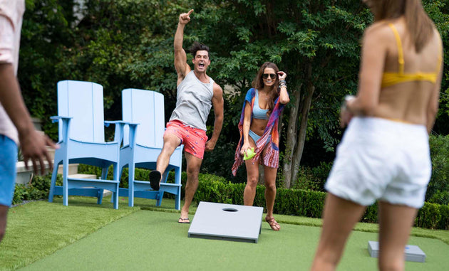 Two people enjoy a lively game of Ledge Lounger Cornhole outdoors. One person celebrates while the other smiles with a drink. Nearby, two others stand by the cornhole set, surrounded by large blue chairs and lush greenery in the background.