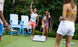 Two people enjoy a lively game of Ledge Lounger Cornhole outdoors. One person celebrates while the other smiles with a drink. Nearby, two others stand by the cornhole set, surrounded by large blue chairs and lush greenery in the background.
