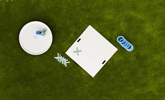 Aerial shot of the Checkers & Tic Tac Toe game by Ledge Lounger on a lawn. A white board with X and O pieces, some askew, is next to a round container full of extras. Blue rings scattered nearby enhance this classic outdoor Tic Tac Toe setup.