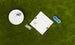 Aerial shot of the Checkers & Tic Tac Toe game by Ledge Lounger on a lawn. A white board with X and O pieces, some askew, is next to a round container full of extras. Blue rings scattered nearby enhance this classic outdoor Tic Tac Toe setup.