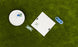 Aerial shot of the Checkers & Tic Tac Toe game by Ledge Lounger on a lawn. A white board with X and O pieces, some askew, is next to a round container full of extras. Blue rings scattered nearby enhance this classic outdoor Tic Tac Toe setup.