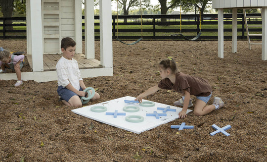 Two children play with Ledge Loungers Checkers & Tic Tac Toe set on the playground amid wood chips. A boy arranges pieces while a girl kneels to place an X. In the background, a child inspects the ground, and swings gently sway nearby.