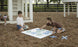 Two children play with Ledge Loungers Checkers & Tic Tac Toe set on the playground amid wood chips. A boy arranges pieces while a girl kneels to place an X. In the background, a child inspects the ground, and swings gently sway nearby.