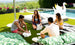 Four people enjoy a playful setting with Dominoes by Ledge Lounger, surrounded by a lawn and greenery. Two relax on leaf-patterned bean bags around a white chess set-topped coffee table. A swan sculpture and pool complete the inviting scene under the clear sky.