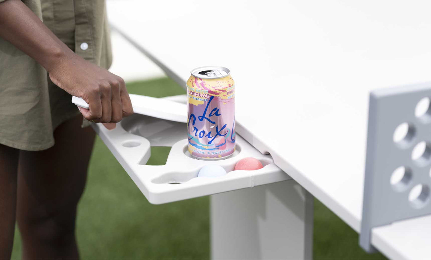 Someone sets a LaCroix can on the white tray from the Ledge Lounger Table Tennis Set outdoors, preparing for entertainment. The tray also holds three ping pong balls, while their other hand grips a paddle against the grassy backdrop.