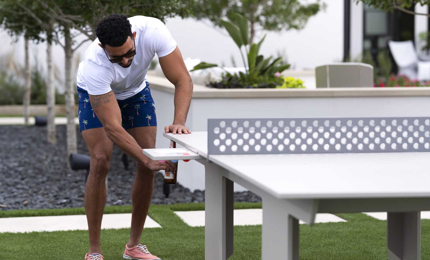 A man in sunglasses and casual summer attire plays on the white Ledge Lounger Table Tennis Set. The scene unfolds in a landscaped yard filled with trees and grass, providing an ideal backdrop for outdoor entertainment.