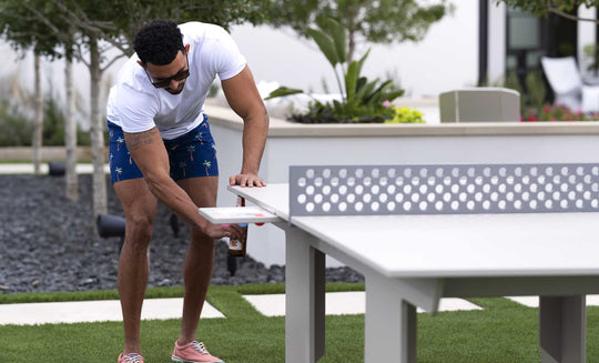 A man in sunglasses and casual summer attire plays on the white Ledge Lounger Table Tennis Set. The scene unfolds in a landscaped yard filled with trees and grass, providing an ideal backdrop for outdoor entertainment.