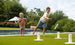 Two children play Ledge Loungers Ring Toss on a grassy lawn by a pool. The older child aims for the stakes as the younger one watches, holding rings. Trees and flowers frame this lively outdoor scene.