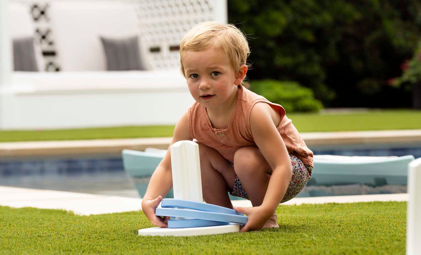 A young child with short blonde hair crouches on grass, playing Ledge Loungers Ring Toss using a blue and white plastic ring. Behind them is a pool and a cushioned seating area amidst greenery. The child sports a sleeveless orange top and patterned shorts—ideal for backyard fun.