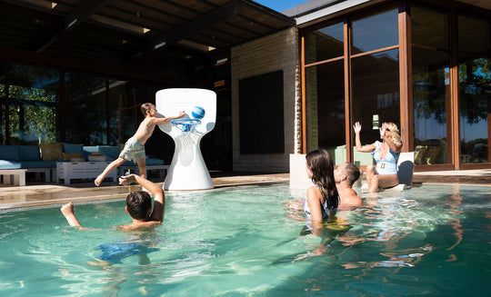 A child leaps off the pools edge, skillfully dunking a basketball into the large Ledge Lounger Hoopstr. Around him, kids and two adults cheer in a lively backyard with modern patio furniture, epitomizing summer fun.