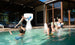 A child leaps off the pools edge, skillfully dunking a basketball into the large Ledge Lounger Hoopstr. Around him, kids and two adults cheer in a lively backyard with modern patio furniture, epitomizing summer fun.