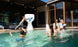 A child leaps off the pools edge, skillfully dunking a basketball into the large Ledge Lounger Hoopstr. Around him, kids and two adults cheer in a lively backyard with modern patio furniture, epitomizing summer fun.