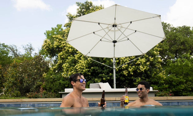 Two people in sunglasses hold beer bottles in a pool, smiling at each other. Behind them is the Ultra Cantilever Umbrella - 13 Octagon by Ledge Lounger, with trees and a cozy patio featuring a white lounge chair.