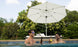 Two people in sunglasses hold beer bottles in a pool, smiling at each other. Behind them is the Ultra Cantilever Umbrella - 13 Octagon by Ledge Lounger, with trees and a cozy patio featuring a white lounge chair.