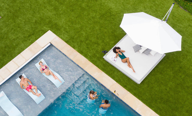 An aerial view reveals a pool scene with two people on floats, a woman relaxing on a sunbed beneath the Ledge Lounger Ultra Cantilever Umbrella - 13 Octagon, and two others chatting in the water. The lush lawn and inviting patio space make it an ideal summer retreat.